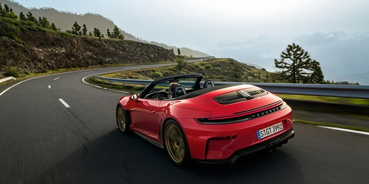 Rear view of red Porsche 911 GT3 RS Cabriolet driving on winding road in scenic landscape.